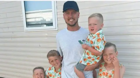 Family photo Nathan with his four children in matching outfits. Two boys with blonde hair to his right and one toddler boy on his hip wear short-sleeved summer shirts with oranges printed on them and green leaves. A girl to his left wears a sleeveless dress in the same print. Nathan is wearing a black cap and light grey t-shirt and they are standing in front of a caravan.