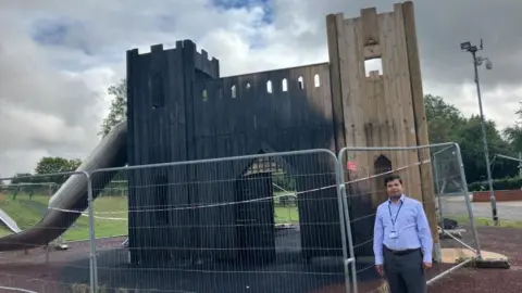 Robbie Kalus/BBC A man stands next to wooden play equipment  