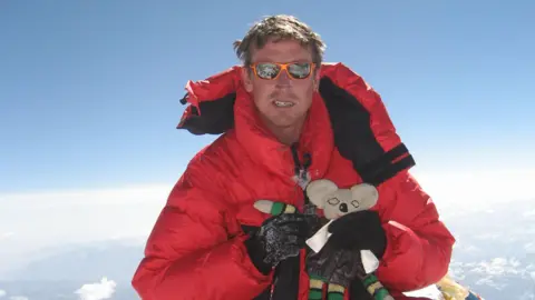 Kenton Cool A man wearing a red puffer jacket and orange sunglasses holds a cuddly toy koala bear as he sits at the summit of Everest. The sky is clear and light clouds can be seen beneath him, sitting on top of other snow-capped mountains.