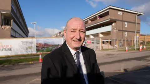 Councillor Steve Evans smiles at the camera while standing in front of the blocks of maisonettes. He wears a black jacket, white shirt and a navy tie.