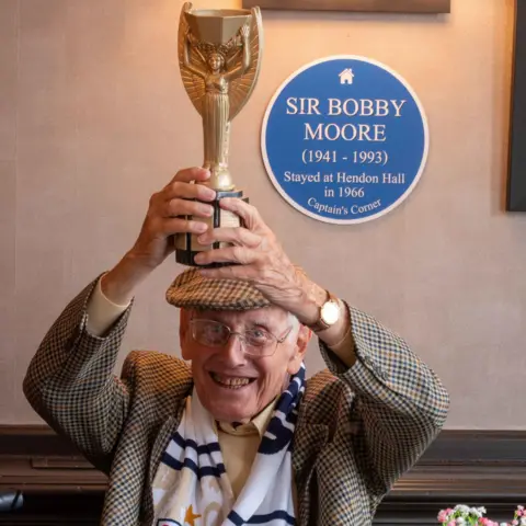 Hendon Hall Care Home Sidney Perez holding up a trophy next to a blue plaque of Bobby Moore