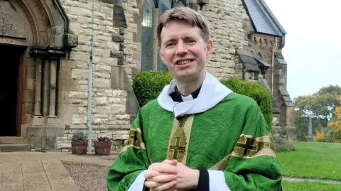 David Chadwick David Chadwick is wearing a green cassock is standing outside his church. He is in his 40s, clean-shaven and has short brown hair with a centre parting. His hands are clasped together in front of him and he is smiling. The church is a large, stone construction with high arch stained glass windows and an ornate arch at the entrance.