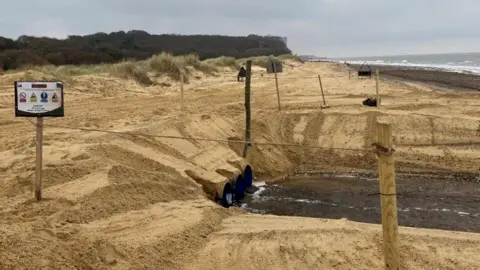 A landscape image of a beach with lots of sand and the ends of three large pipes coming out from underneath. The sea is in the distance to the right of the image and on the left there are trees and buses. 