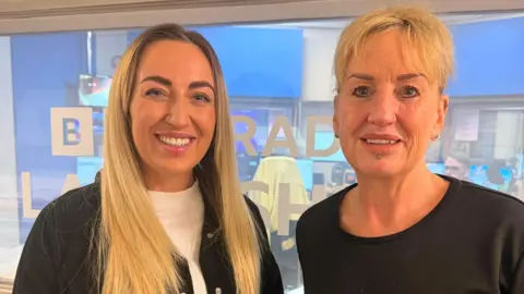 Becky Hobkirk has long blonde hair a white sweater and black jacket. Mum Nicky has shorter blonde hair and a black top. Both are smiling in front of BBC Radio Lancashire glass door. 