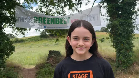 Charlotte is standing in front of a sign saying "this is green belt not grey" and a tree to the left and to the right of the sign.