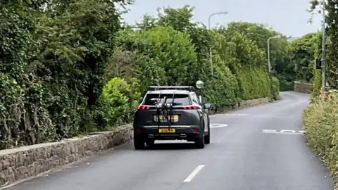 Route de Noirmont. A narrow road with trees and bushes either side. The picture shows the white lines running along the road until they stop and don't go any further. 