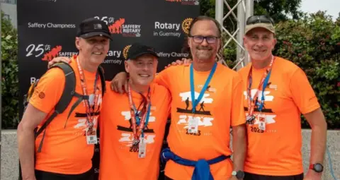 Four men in bright orange T-shirts with a medal around their works posing for a picture in front of a black board that has branding including Rotary of Guernsey's logo.