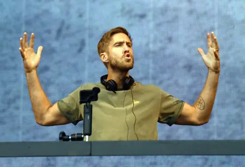 PA Media A man in a green T-shirt with black headphones around his neck. He is standing behind a desk and has his arms in the air. 
