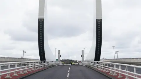 A view of the single-carriageway road that carries traffic in both directions over the Gull Wing bridge. There are barriers either side of the road and a pavement beyond them. Two large white and black "wings" point upwards from the middle of the bridge.