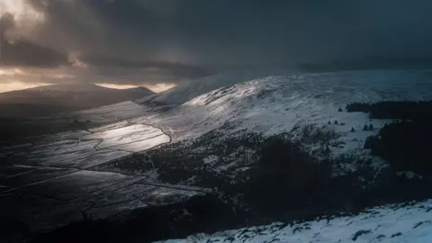 Adam Morgan/Caira Kaneen Picture taken atop a hill on the Isle of Man on a snowy, dark day. Light can be seen breaking through the clouds onto the snowy land.