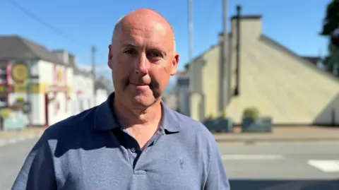 The image shows a middle-aged bald man standing outdoors on a street on a sunny day. He is wearing a blue polo shirt and looking straight at the camera.
Buildings and trees visible in the background.