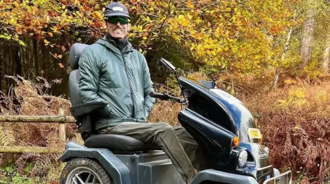 Tom Staniford Tom Staniford is smiling as he sits in a mobility scooter outside with bracken and a beech hedge behind him. He is wearing a cap, dark green sunglasses, a scarf, anorak and waterproof trousers.