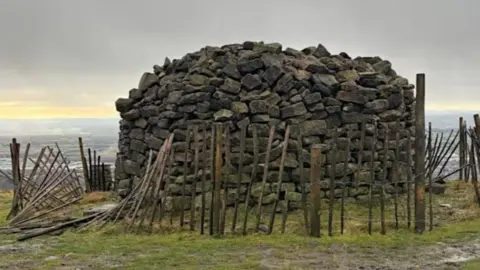 Large hut-size cairn made of large stones and surrounded by a circle of wooden fencing, which has partially fallen. It stands at the top of a hill, with flatter land spread out below.
