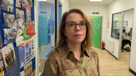 Andrea Cleaver stands in a bright corridor in the Welsh Refugee Council offices, beside a colourful bulletin board covered with photos and paper crafts. Green doors and office spaces are visible further down the hallway. She has shoulder-length light brown hair and wear glasses and pink lipstick. 