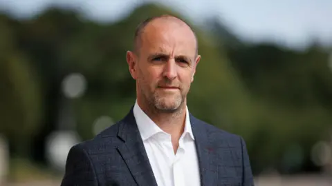 PA Media Mark H Durkan in a blue checked jacket and open neck white shirt. He has stubble and his head is mostly shaved.