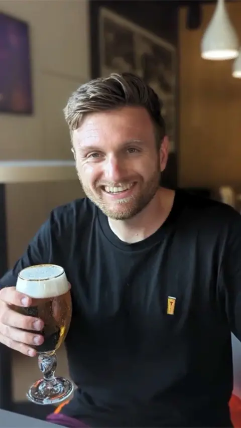 A head-and-shoulders image of Harry Oates. He is looking straight at the camera and smiling. He has light brown hair, longer at the top and shaved at the sides and a trimmed beard. He has blue eyes and is dressed in black t-shirt.