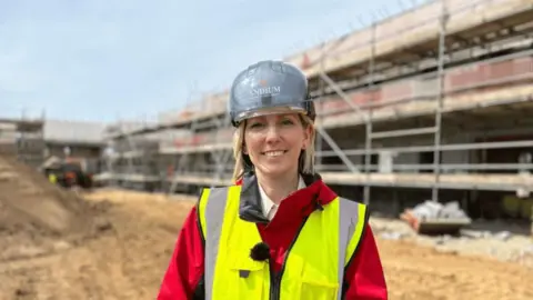 New Andium CEO Lindsay Wood stands on a building site. Scaffolding is visible behind her. She is wearing a red coat with a fluorescent vest and a helmet. 