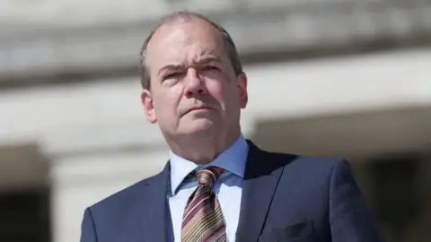 PA Media Sir Michael McBride - he has dark hair and is wearing a navy suit, white shirt and red, gold and black tie.