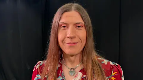 Ben Schofield/BBC Jessica Brown looking directly at the camera. She has straight brown hair, which falls just below her shoulders. She is wearing a red dress with large flowers on it, though only her head and shoulders are in the frame. Around her neck is a silver chain with a heart-shaped padlock pendant. She is smiling at the camera and is sitting in front of a black backdrop.