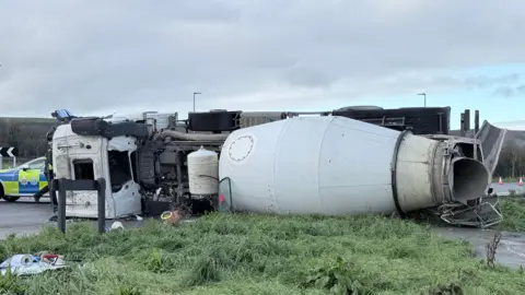 The image shows a close-up of the overturned HGV lying on the grass, with a police car visible in the back on the left-hand-side. The sky is a light blue with grey clouds.
