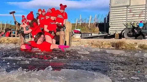 January Daily Dip A group of people in red t-shirts are coming down steps to get into a pond of frozen water, there is a sauna in the background. 