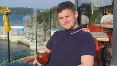 Ed O'Brien wearing a navy top and holding a glass, with ocean behind him.