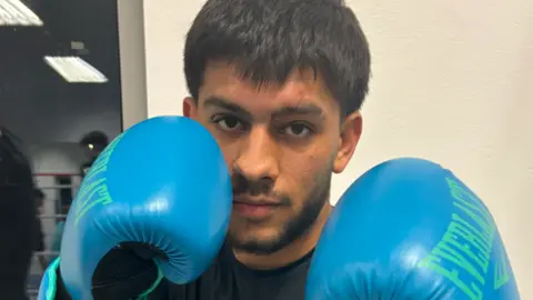 BBC Shows young man with a dark hair and a beard in a dark T-shirt and light blue boxing gloves 