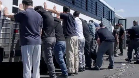 EPA/US Immigration and Customs Enforcement handout A still frame from a video shows a group of men, with their backs to the camera and hands on the side of a white coach with black bars on its windows during an immigration raid at the Hyundai-LG vehicle assembly plant in Ellabell, Georgia.
The men are in casual clothing, mostly jeans or slacks and T-shirts.