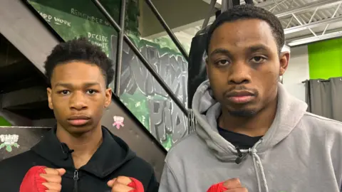 Bryce and Maurice stand with their hands up in boxing pose. Bryce has short black hair and is wearing a black hoodie, Maurice has short black hair and is wearing a light grey hoody.