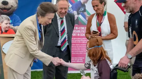 Princess Anne, the Princess Royal makes a presentation to a player in a wheelchair at Tag Fest 26. The Princess is wearing as a buff-coloured trouser suit over a blue blouse. The player shaking hands with her is in a wheelchair. They have red hair and glasses that are held on with a plastic band around their head.