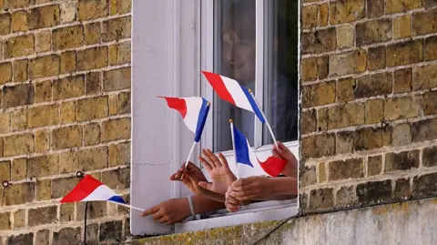 AFP via Getty Images A young boy is seen behind a window which has his hands and the hands of other young people sticking out of a gap at the bottom of it while waving the red, white and blue flag of France.