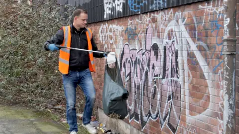 Man in white pumps, blue jeans, orange tabard and black jacket collecting rubbish with a litter grabber next to a wall covered in graffiti