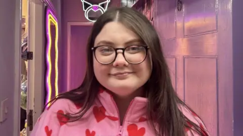 A woman with long brown hair and glasses in a pink top with hearts on, standing in a corridor lit with a pink light