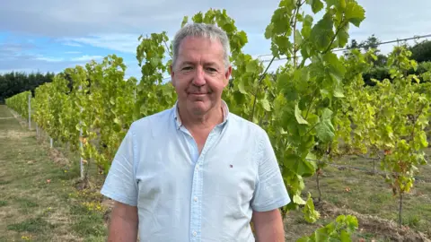 Tim Crowley sonríe a la cámara. Está de pie frente a un viñedo y lleva una camisa azul claro.
