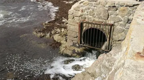 Storm overflow pipe at a beach in Cornwall. The pipe is coming from beneath a platform overlooking the sea. The hole is covered by a metal grate and liquid is pouring out of the pipe and into the sea