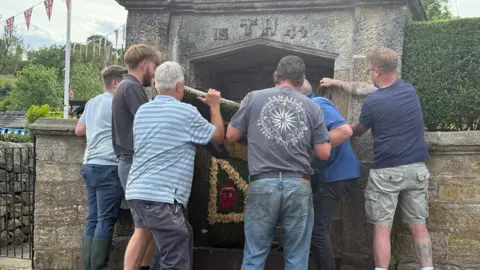 A group of people lift a decorative board onto a stone well. There is a gate to the side and a hedge on the other side of the well.