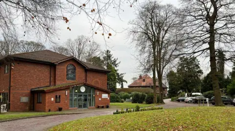 St Joseph's Hospice The main entrance to the hospice, with a large glass panelled entrance and a stained glass window above. Grassed areas and trees can also be seen all around, with another building and parked cars seen in the distance.