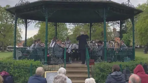 Martin Heath/BBC The bandstand with green poles and railings and a black roof. There is a band dressed in white shirts and dark trousers playing various brass and percussion instruments. There is a low hedge in front of the bandstand and stone steps leading down from the raised platform to the ground. A number of people are standing in front of the bandstand listening to the music.