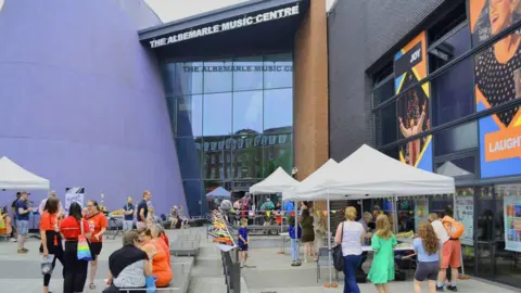 Hull Truck Theatre A tall building with "The Albemarle Music Centre" lettering across the top, It has a purple wall on the left and there are large glass panes down the middle. In front of the building is an outside area with grey seats and marquees and bunting. People are sitting down and wandering around.