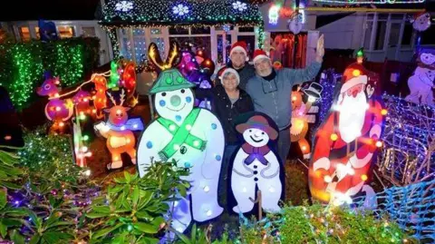 A house covered in multi-colour Christmas lights with a family of three, mum, dad and son, standing in front of the house. They are surrounded by illuminated Christmas figurines, including a two snowmen and Santa. 