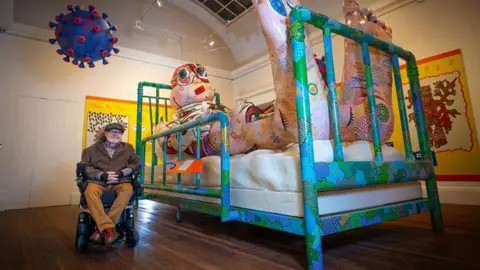 Artist Jason Wilsher-Mills sits alongside one of his exhibits - a giant inflatable character lying in a hospital bed watching TV inside the Grundy Art Gallery.