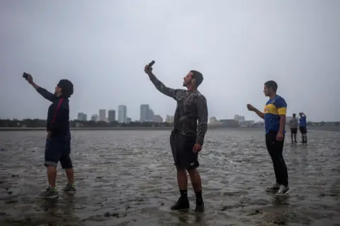 Reuters The Tampa skyline is seen in the background as local residents (L-R) Rony Ordonez, Jean Dejesus and Henry Gallego take photographs after walking into Hillsborough Bay ahead of Hurricane Irma in Tampa, Florida, 10 September