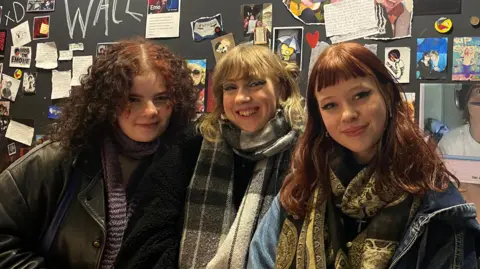 Three women - from near  - Maddie, Seren and Molly lasting  successful  beforehand   of a show  of tiny  pictures and notes. Maddie has dark, curly shoulder-length hairsbreadth  and is wearing a scarf and leather jacket. Seren has acheronian  blonde hair, with a checked scarf and overgarment  and Molly has reddish  hair, fringe and wears a denim overgarment   and paisley scarf.