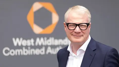West Midlands Combined Authority A man with light grey hair, wearing black glasses, a white shirt and black suit jacket, pictured from the shoulders up in front of a grey background with an orange logo and the words "West Midlands Combined Authority".