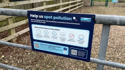 A blue sign on a metal gate says "help us spot pollution" and lists signs that water may be polluted, including dead fish or odours of sewage
