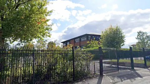 Google A school building set against bright skies is behind high black metal fencing. There are bushes and trees in the grounds of the school.