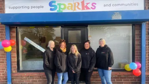 Five women wearing Sparks hoodies stand outside their new centre in Eastfield, with the Sparks name above the door and coloured balloons on either side.