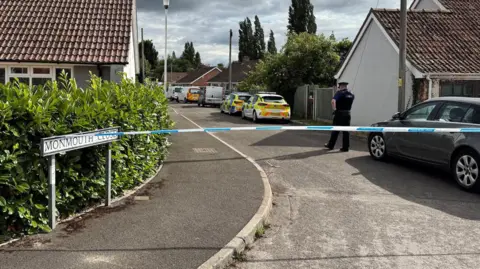 A street closed off by blue and white police tape. There is a grey sky with a police officer walking away behind the tape.