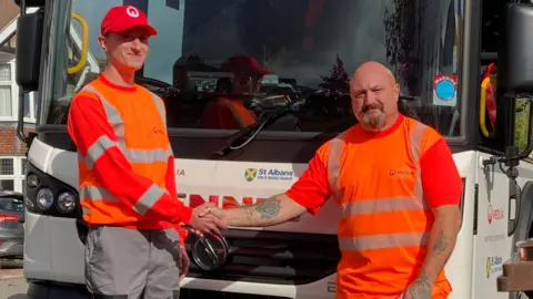 BBC/Justin Dealey Two refuse collectors dressed in orange are stood next to a white St Albans bin lorry. they are shaking hands.