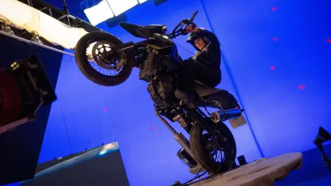 Alamy Cameron Diaz pulls a wheelie on a black motorcycle against a blue background inside a film studio.